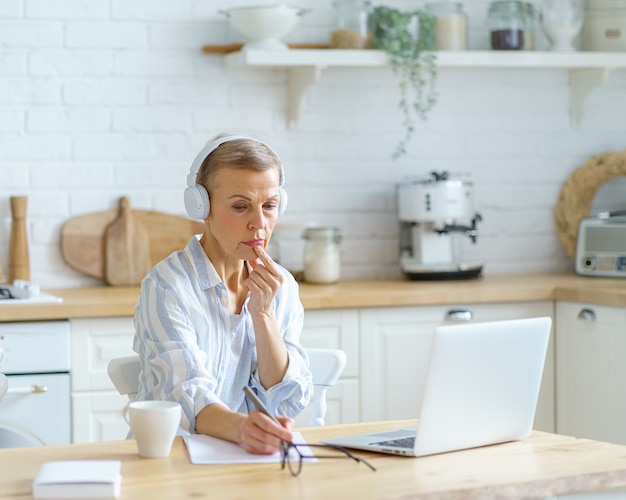 Mujer de mediana edad usando audífonos tomando notas mientras estudia en línea en la cocina