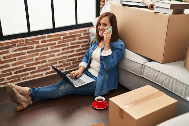 Mujer de mediana edad usando una computadora portátil y hablando por teléfono inteligente en un nuevo hogar