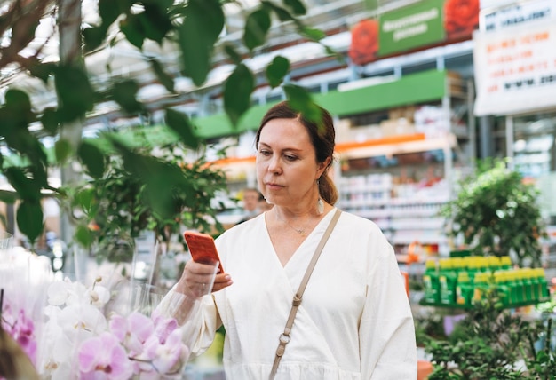 Mujer de mediana edad con vestido blanco toma una foto en el móvil de plantas de interior en macetas verdes en la tienda de jardinería