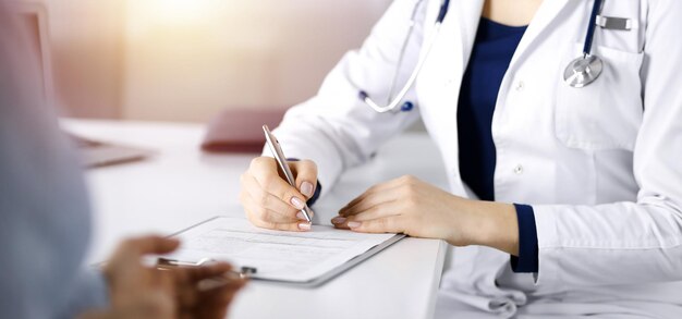 Una mujer médica desconocida está prescribiendo algunos antibióticos para su paciente, mientras están sentados en el escritorio en el gabinete soleado de una clínica.