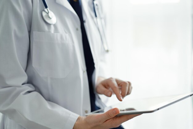 Mujer médica usando una tableta mientras está de pie cerca de una ventana panorámica en la clínica, de cerca. Médico o cirujano en el trabajo. Concepto de medicina