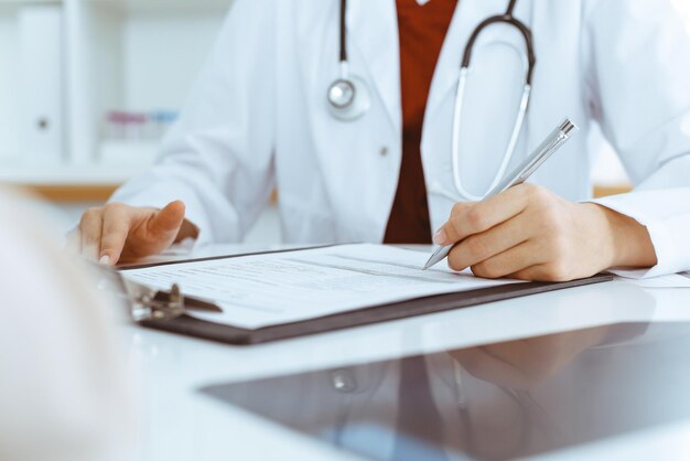 Una mujer-médico desconocida llenando un formulario de solicitud mientras consulta a un paciente.