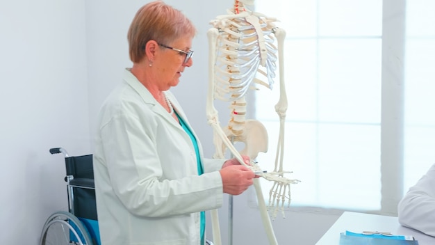 Mujer médico enseñando anatomía con modelo de esqueleto humano de pie en la oficina de conferencias del hospital. Terapeuta experto de la clínica hablando con colegas sobre la enfermedad, profesional de la medicina.