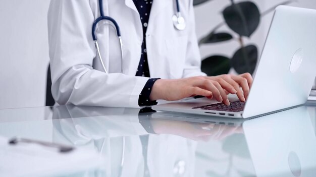 La mujer médico está escribiendo en una computadora portátil en una mesa de vidrio en la clínica de medicina de vista horizontal y