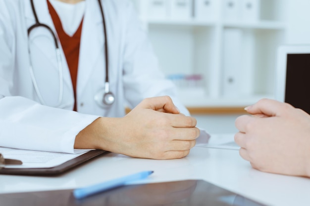 Mujer-médico y paciente desconocidos discutiendo algo mientras están sentados en la mesa en la clínica, en primer plano. Concepto de medicina.