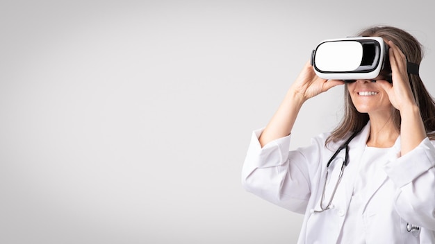 Mujer médico senior con gafas de realidad virtual de pie en el panorama de fondo de estudio gris con