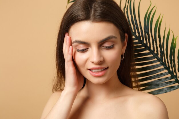 Mujer medio desnuda morena de cerca con una piel perfecta, maquillaje desnudo y hoja de palma aislada en la pared de color beige pastel