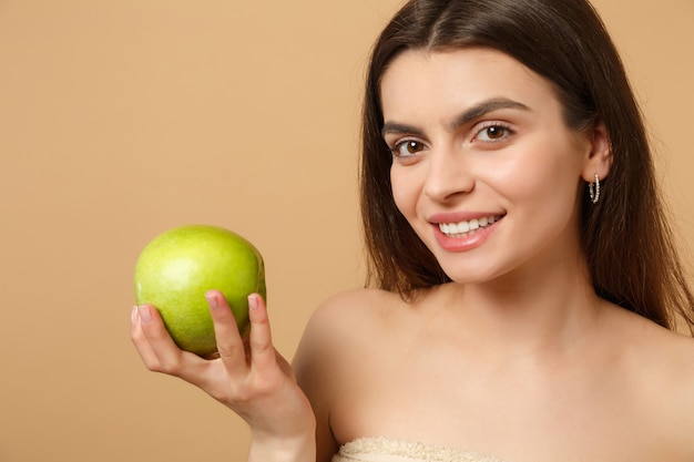 Mujer medio desnuda morena de cerca con una piel perfecta, maquillaje desnudo sostiene manzana aislada en la pared de color beige pastel