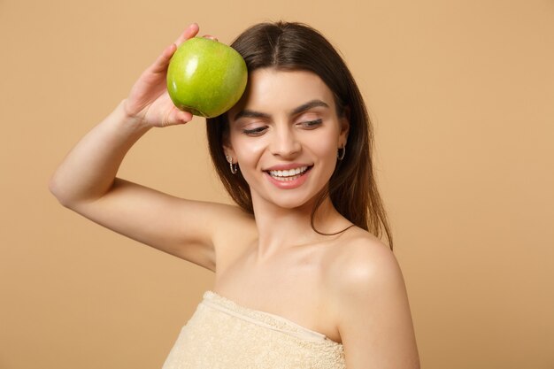 Mujer medio desnuda morena de cerca con una piel perfecta, maquillaje desnudo sostiene manzana aislada en la pared de color beige pastel