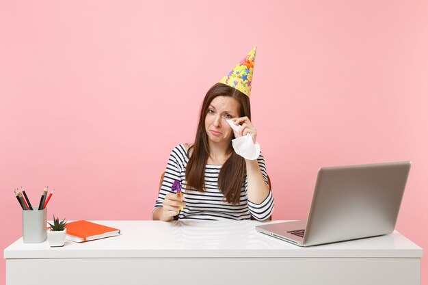 Mujer melancólica con sombrero de fiesta llorando secándose las lágrimas con un pañuelo porque celebrando el cumpleaños solo en el trabajo en el escritorio blanco con un portátil aislado sobre fondo rosa. Logro de carrera empresarial. Copie el espacio.