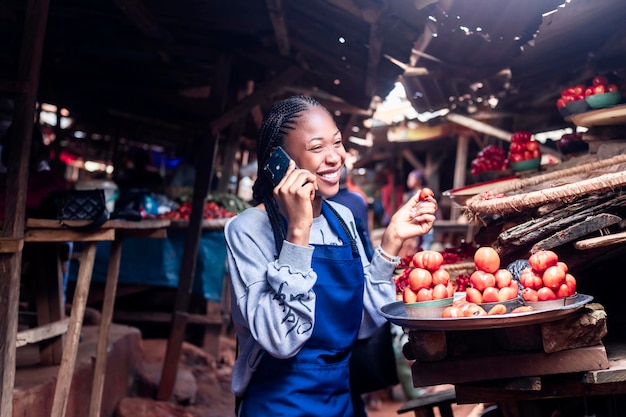 Mujer del mercado africano en delantal azul vendiendo artículos haciendo una llamada telefónica en un mercado