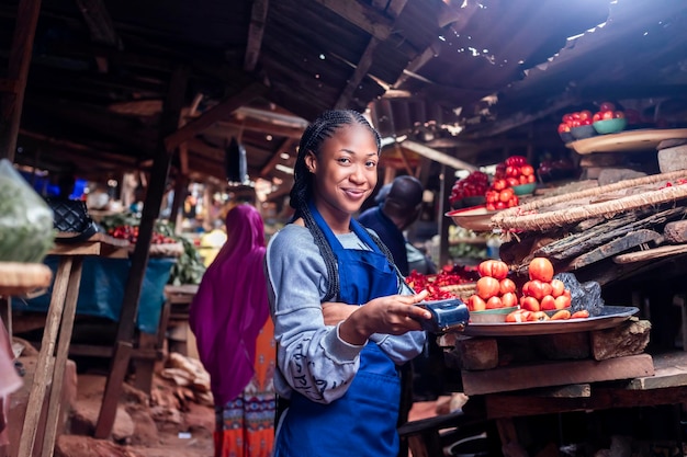 Mujer del mercado usando una terminal pos para comprar y vender en un mercado local típico de tomates y verduras africanos