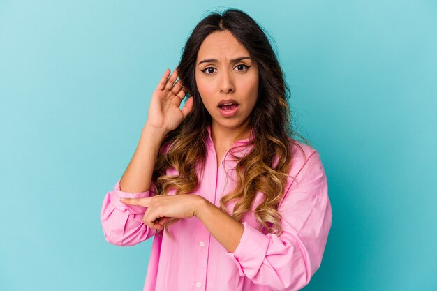 Mujer mexicana joven aislada en la pared azul que intenta escuchar un chisme.