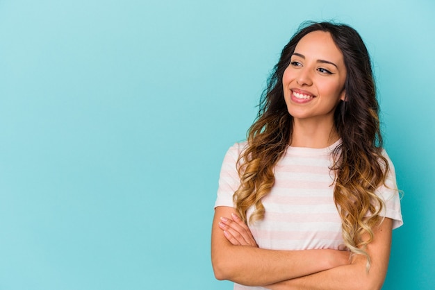 Mujer mexicana joven aislada en la pared azul sonriendo confiada con los brazos cruzados.