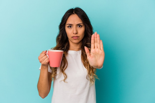 Mujer mexicana joven que sostiene una taza aislada en azul que se coloca con la mano extendida que muestra la señal de stop, que le impide.