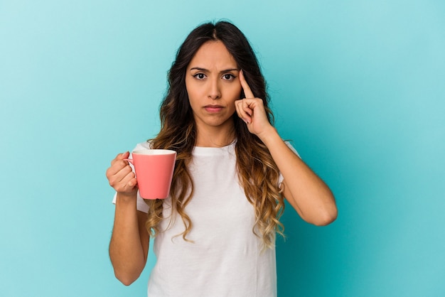 Mujer mexicana joven sosteniendo una taza aislada sobre fondo azul señalando el templo con el dedo, pensando, centrado en una tarea.