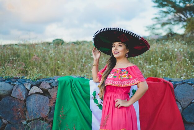 Foto mujer mexicana con sombrero y vestido tradicionales junto a la bandera mexicana retrato al aire libre