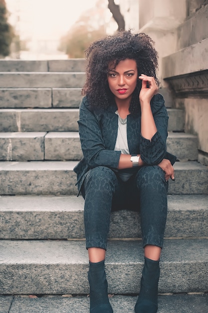 Mujer mezclada joven con el peinado afro que sonríe en fondo urbano. Chica negra con ropa casual.