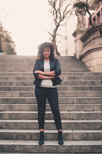 Mujer mezclada joven con el peinado afro que sonríe en fondo urbano. Chica negra con ropa casual.