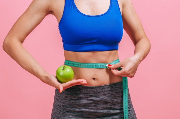 La mujer mide la cintura y muestra una manzana verde en una pared rosa.