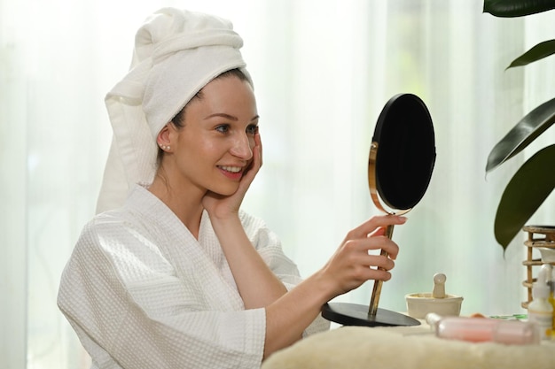 Mujer milenaria sonriente en bata de baño parada frente al espejo y haciendo rutina diaria de belleza en casa