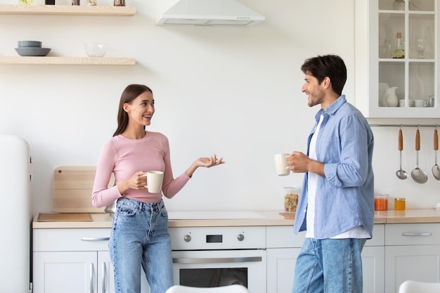 Mujer milenaria sonriente satisfecha y chico con tazas de bebida caliente desayunan hablando en el interior de la cocina