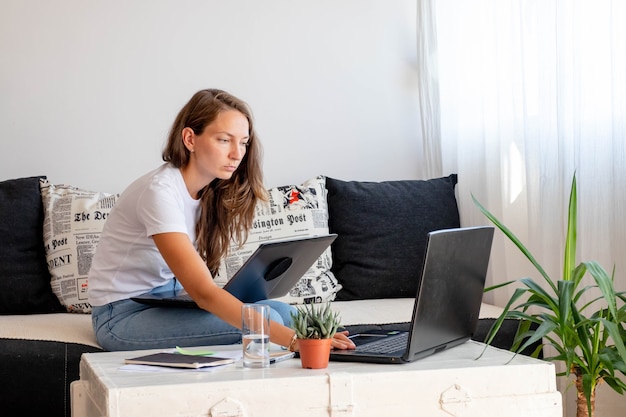 Mujer milenaria trabaja en casa, asientos cerca de la computadora portátil con portátil, vaso de agua en la mesa blanca en el espacio de trabajo doméstico.
