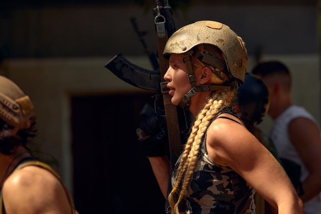 Mujer militar caucásica con equipo táctico posando para una foto en la temporada de verano con uniforme de camuflaje verde y rifle de asalto con equipo militar y la dama de los auriculares está mirando al lado