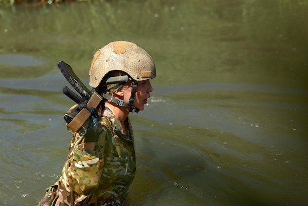 Mujer militar caucásica con equipo táctico posando para una foto en la temporada de verano con uniforme de camuflaje verde y rifle de asalto con equipo militar y la dama de los auriculares está mirando al lado