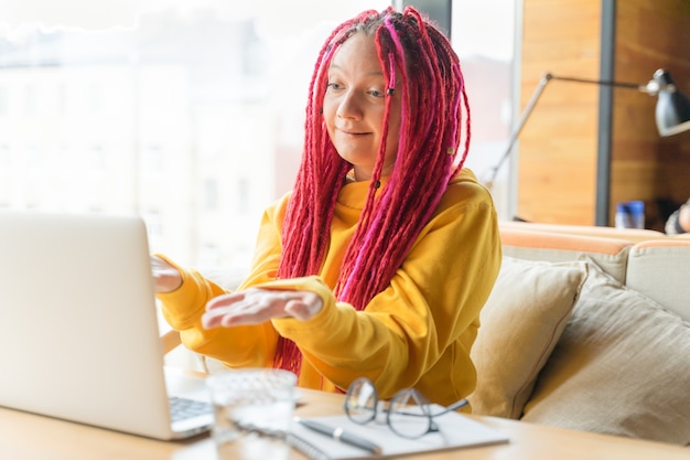 La mujer mira la computadora portátil y se sorprende, indignada, extiende sus manos. Chica con largo cabello rosado