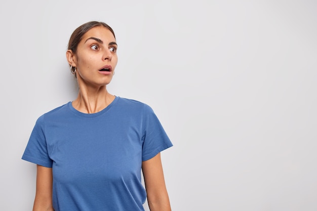 Mujer mira fijamente los ojos saltones ha contenido el aliento vestido con una camiseta informal no puede creer en algo aterrador en el espacio de copia en blanco para promo