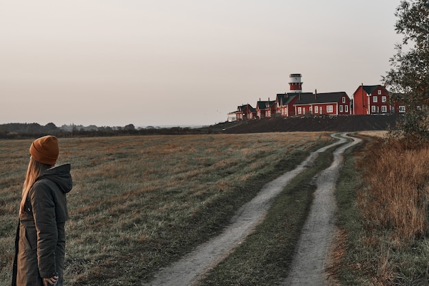 Una mujer mira a lo lejos de un camino rural, un faro rojo y blanco al final del camino. Viajar, amanecer.