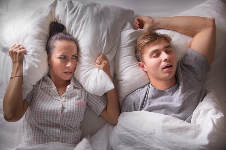 La mujer mira a su novio molesto sosteniendo una almohada sobre sus orejas mientras ronca acostado de espaldas con la boca abierta