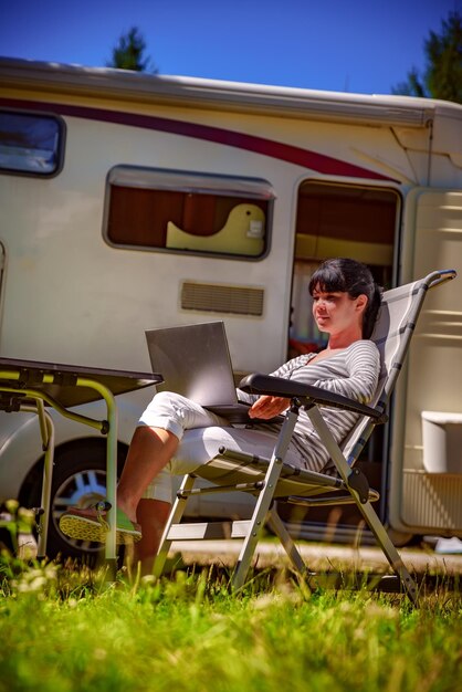 Mujer mirando la computadora portátil cerca del campamento. Vacaciones en coche caravana. Viajes de vacaciones en familia, viaje de vacaciones en autocaravana. Tecnología de comunicación de información de conexión Wi-Fi.