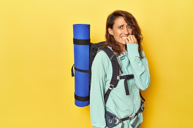 Mujer con mochila de senderismo y colchoneta en amarillo mordiéndose las uñas nerviosa y muy ansiosa