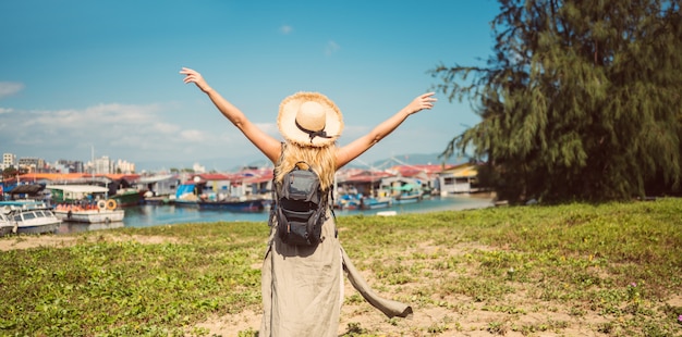 La mujer del mochilero rubio del viajero en la opinión trasera del sombrero de paja camina a lo largo de la costa en los barcos de pesca del embarcadero. Viaje de aventura en China. Isla tropical de Asia turístico. Concepto de viaje de vacaciones de vacaciones de verano