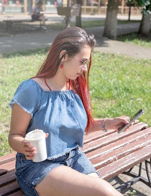 Mujer de moda alegre con cabello rojo bebiendo café en el parque tomando selfie