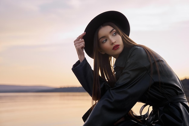 Mujer de moda de belleza en impermeable de cuero y sombrero negro redondo cerca del estanque del lago. Retrato de niña al atardecer. Hermoso maquillaje y lápiz labial rojo brillante en los labios