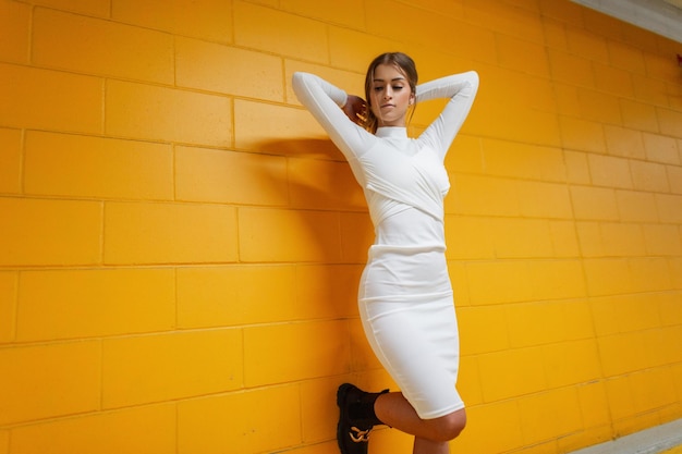 Mujer de moda bonita sexy con cuerpo esbelto en vestido blanco de moda posa cerca de una pared de ladrillo amarillo de color en el interior