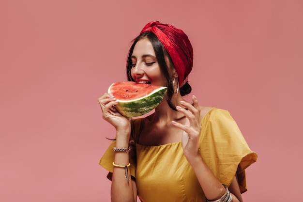 Mujer de moda con cabello moreno en aretes de diadema roja y ropa brillante comiendo sandía en un fondo aislado