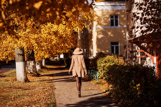 La mujer de moda se está yendo por la calle en otoño. La mujer lleva un sombrero de ala ancha y un abrigo beige.