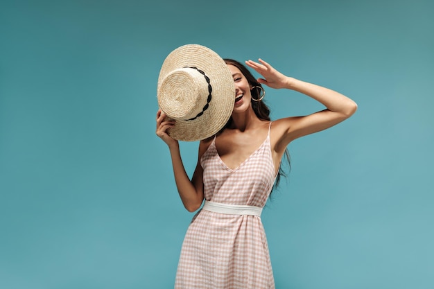 Mujer de moda feliz con cabello largo y negro en aretes vestido fresco con sombrero moderno y riendo en un fondo aislado