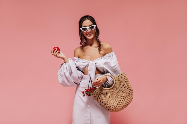 Mujer de moda con gafas de sol blancas blusa moderna falda larga a rayas posando con una linda sonrisa y sosteniendo una manzana roja y una bolsa fresca con flores