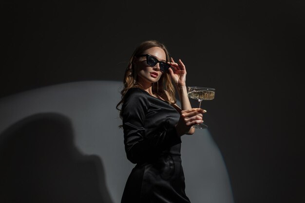 La mujer de moda, hermosa y elegante, usa gafas de sol geniales con un vestido negro de noche de moda con una copa de champán en una fiesta, una dama elegante y bonita celebra un evento