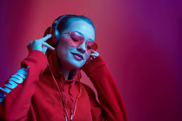 La mujer de moda hipster usa elegantes gafas y auriculares escuchando música sobre fondo de color neón en el estudio.