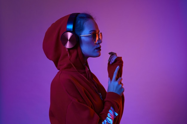 La mujer de moda hipster usa elegantes gafas y auriculares escuchando música sobre fondo de color neón en el estudio.