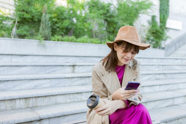 Mujer de moda joven vestida con ropa de otoño se sienta en un banco y usa un teléfono inteligente al aire libre