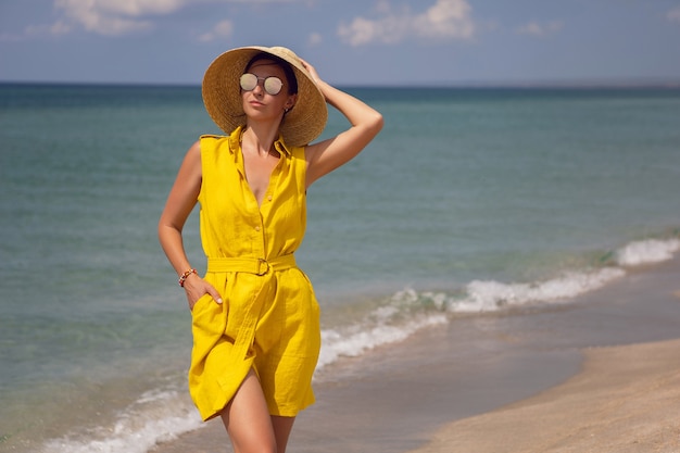 Mujer de moda joven con un vestido de sombrero amarillo y gafas de sol está posando en la playa en verano de vacaciones
