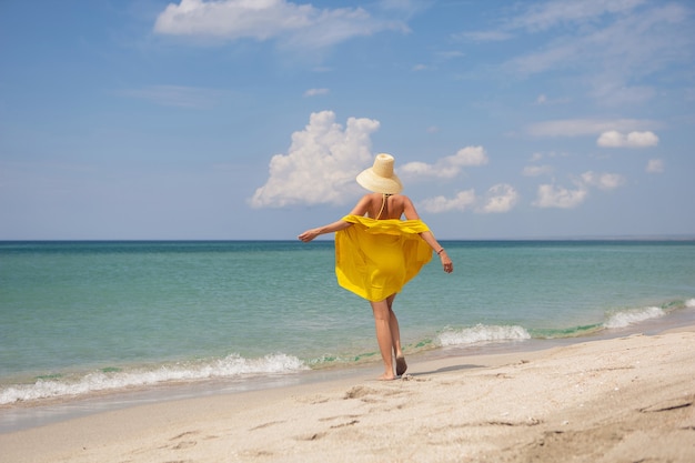 Mujer de moda joven con un vestido de sombrero amarillo y gafas de sol está posando en la playa en verano de vacaciones