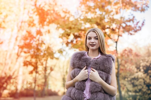 Mujer de moda. Niña sonriente en posin de abrigo de piel en otoño parque con árboles y hiedra